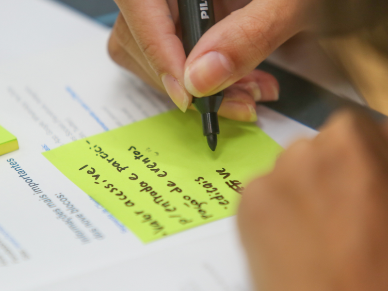 A imagem é uma fotografia em que aparece uma mão escrevendo com uma caneta preta em um post-it. O post-it está em cima de um modelo de negócios impresso. A imagem está ilustrando a atividade de modelagem de negócios do Desafio Unicamp 2024. Fim da descrição.