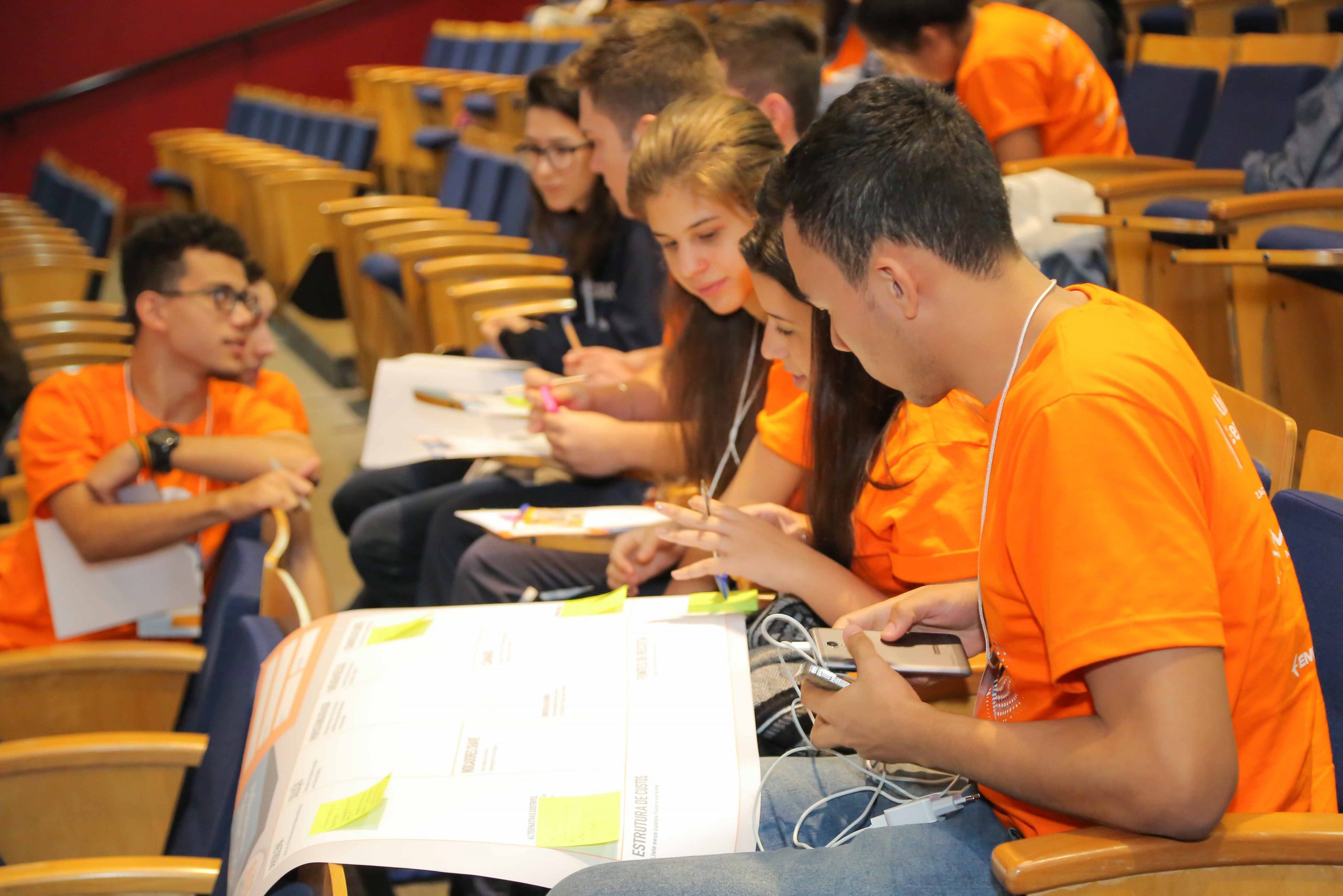 jovens sentados em um auditório com um cartaz no colo. O cartaz tem papéis colados e os jovens estão com canetas na mão e vestindo camiseta laranja.