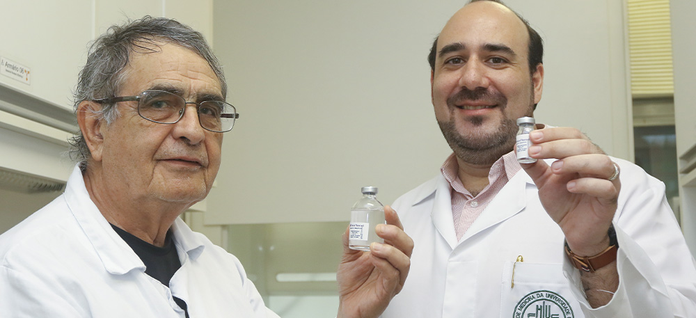 Foto de dois pesquisadores homens brancos que seguram um frasco do remédio oncotherad. Ambos vestem jaleco branco, um é mais velho, usa óculos e tem cabelos brancos e o outro é mais novo, calvo com barba preta. Fim da descrição.