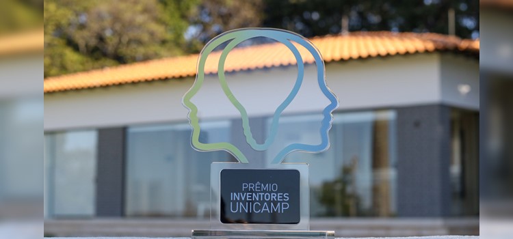 Foto de um troféu em foco do Prêmio Inventores da Unicamp. O troféu é o logo do Prêmio, que forma uma lâmpada com duas metades de cabeça ao redor. Ele é em degradê do verde ao azul como as cores da Inova Unicamp. Ao fundo o prédio da Inova Unicamp desfocado. Fim da descrição.