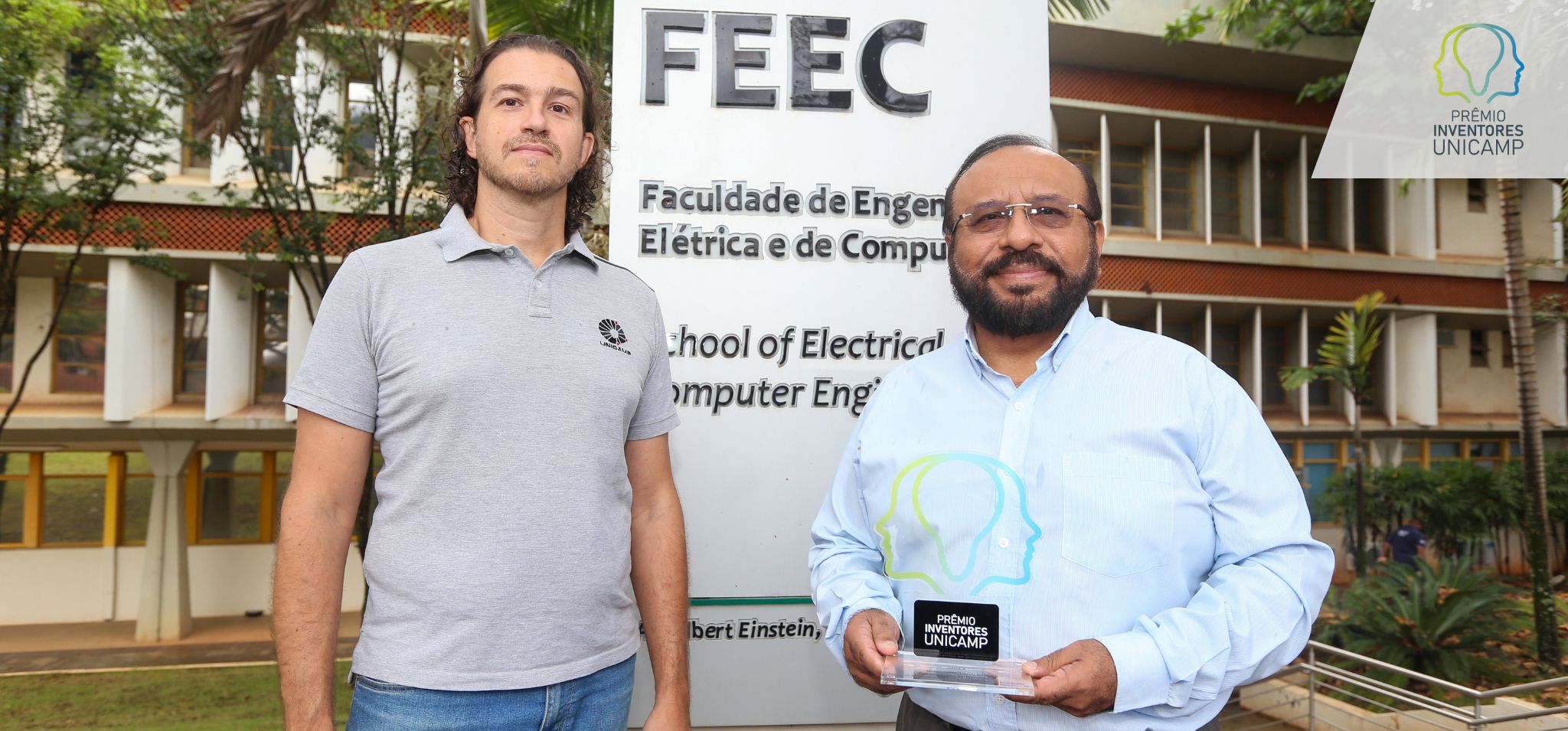 Homem de camisa cinza (a esquerda) e homem de branco (a direita) segurando a premiação em frente a placa da FEEC