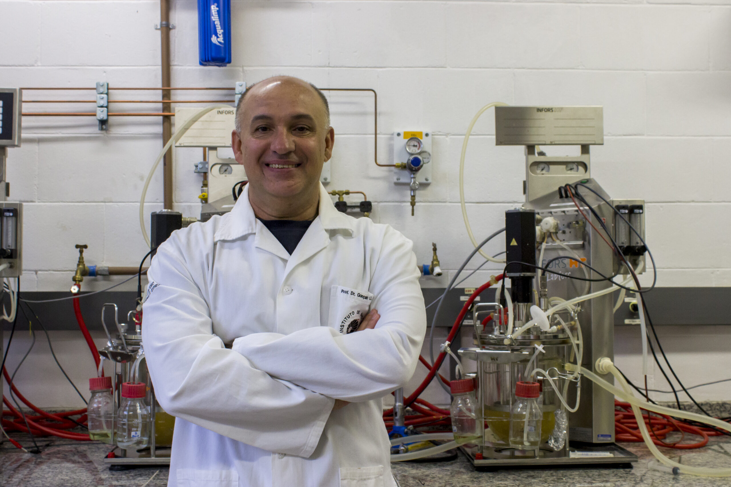 Prof. Gonçalo de jaleco, sorrindo em frente à equipamentos do laboratório