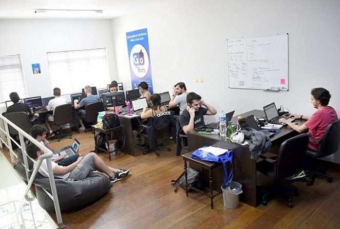 foto mostra o escritório da empresa gobots com funcionários sentados em escrivaninhas trabalhando em laptops e outros sentados em pufes. O ambiente é descontraído