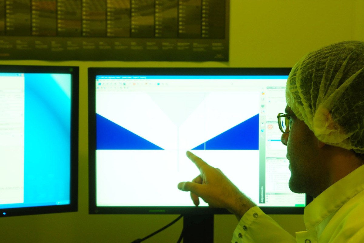 A foto está com luz baixa amarelada e mostra parte de um homem branco, de óculos, usando jaleco e touca, apontando para um resultado em uma das duas telas de computador a sua frente. Não é possível ler o que está na tela.