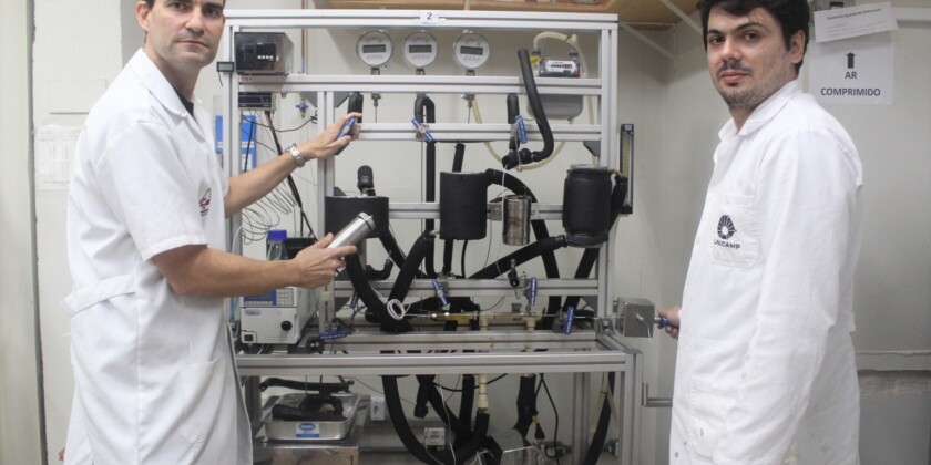 Dois homens, pesquisadores da Unicamp, vestindo jaleco branco, aparecem em frente ao equipamento usado na produção de aromas naturais de frutas. A máquina tem estrutura metálica, da altura dos cientistas, além de tubos pretos e mostradores digitais.