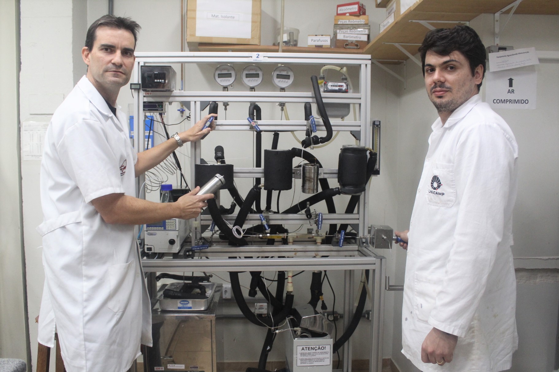 Dois homens, pesquisadores da Unicamp, vestindo jaleco branco, aparecem em frente ao equipamento usado na produção de aromas naturais de frutas. A máquina tem estrutura metálica, da altura dos cientistas, além de tubos pretos e mostradores digitais.