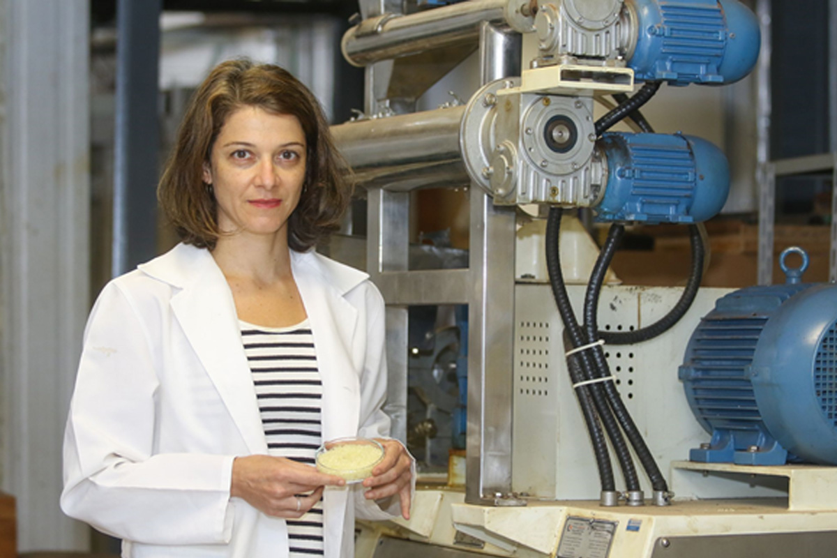 A foto é da pesquisadora Ana Silvia Prata, que está vestindo um jaleco branco em cima de uma blusa listrada. Ela tem cabelo curto na cor castanho e está segurando um material referente a tecnologia. Atrás dela, no canto direito, possui máquinas de laboratório.