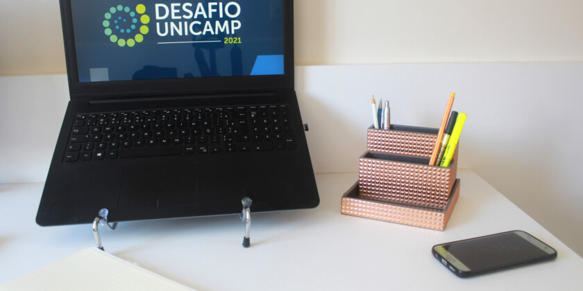 Mesa com computador escrito Final Desafio Unicamp, caderno, porta-lápis e celular