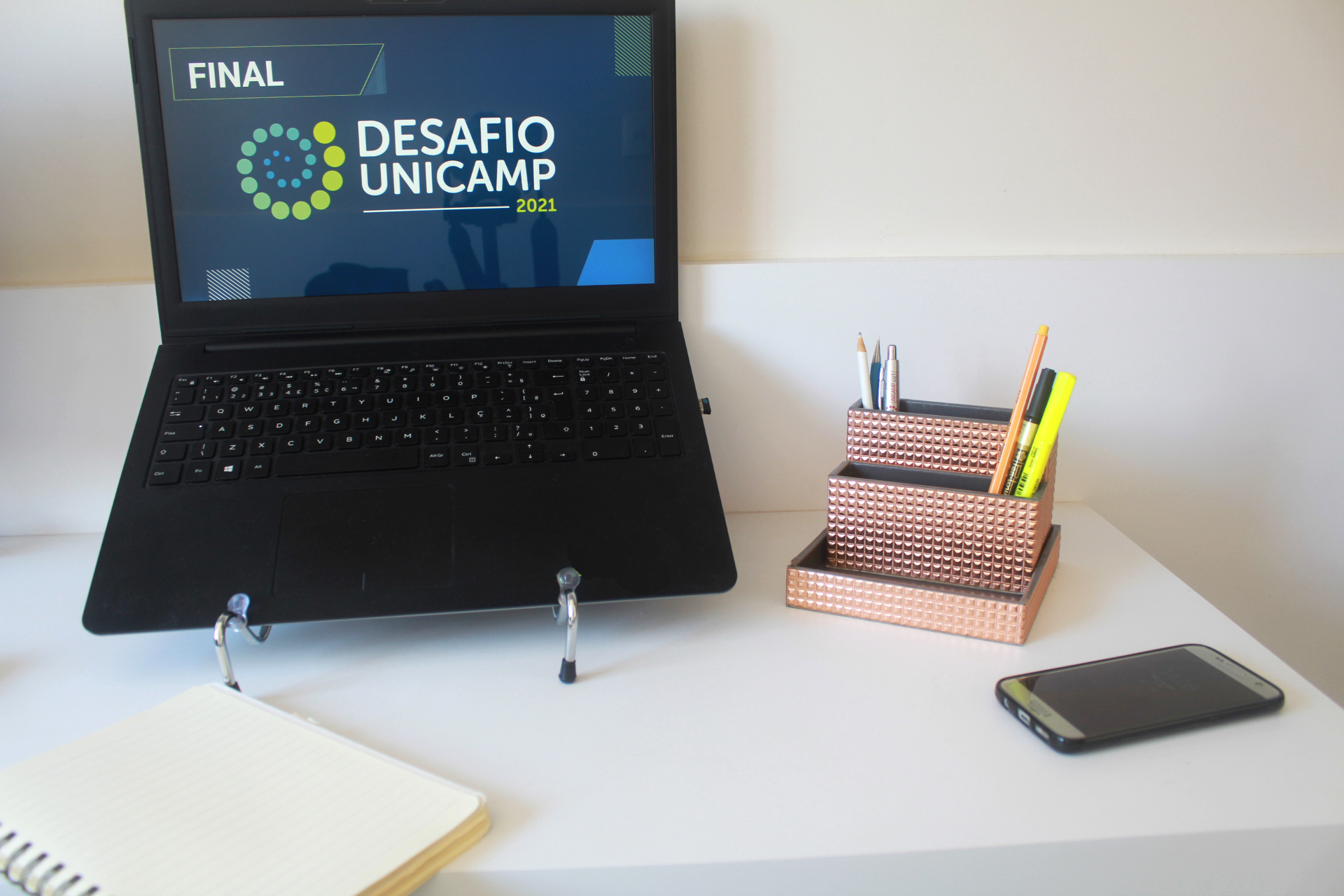 Mesa com computador escrito Final Desafio Unicamp, caderno, porta-lápis e celular
