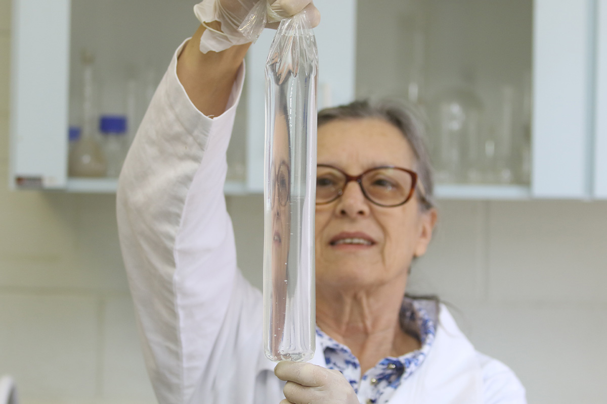 Mulher idosa com óculos e jaleco está em foco, segurando com as duas mãos um plástico em formato vertical e com um líquido transparente. Atrás dela está um armário com tudos de ensaio.
