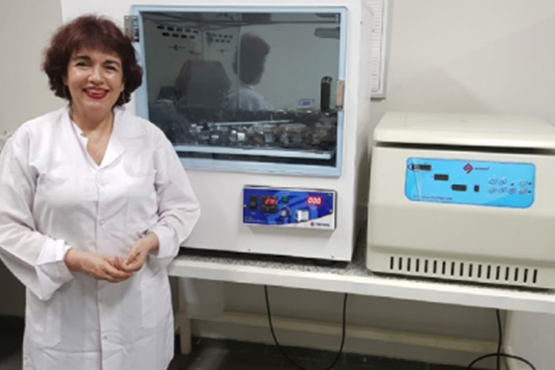 Mulher na faixa dos 55 anos está com um jaleco e ao lado de máquinas em laboratório. Ela tem cabelo castanho e está sorrindo.