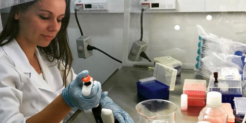 Professora Catia Ornelas em um laboratório de pesquisa. A professora veste um jaleco branco e possui cabelos escuros. Ela está manuseando materiais de pesquisa.