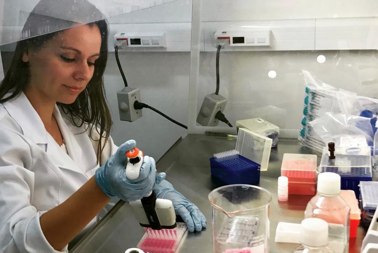 Professora Catia Ornelas em um laboratório de pesquisa. A professora veste um jaleco branco e possui cabelos escuros. Ela está manuseando materiais de pesquisa.