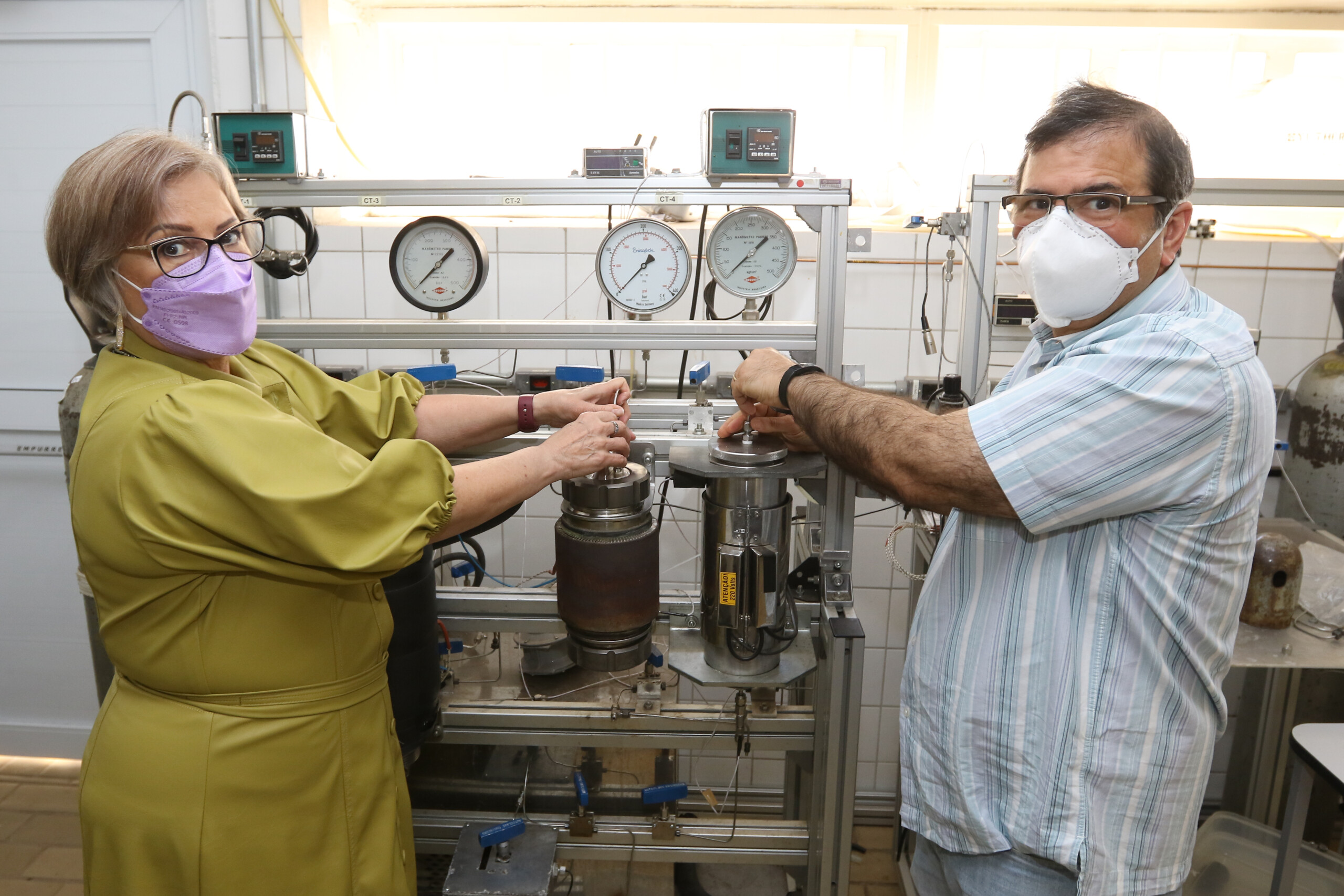 Os pesquisadores estão mexendo no aparelho de extração supercrítica dentro do laboratório, ambos estão de máscara.