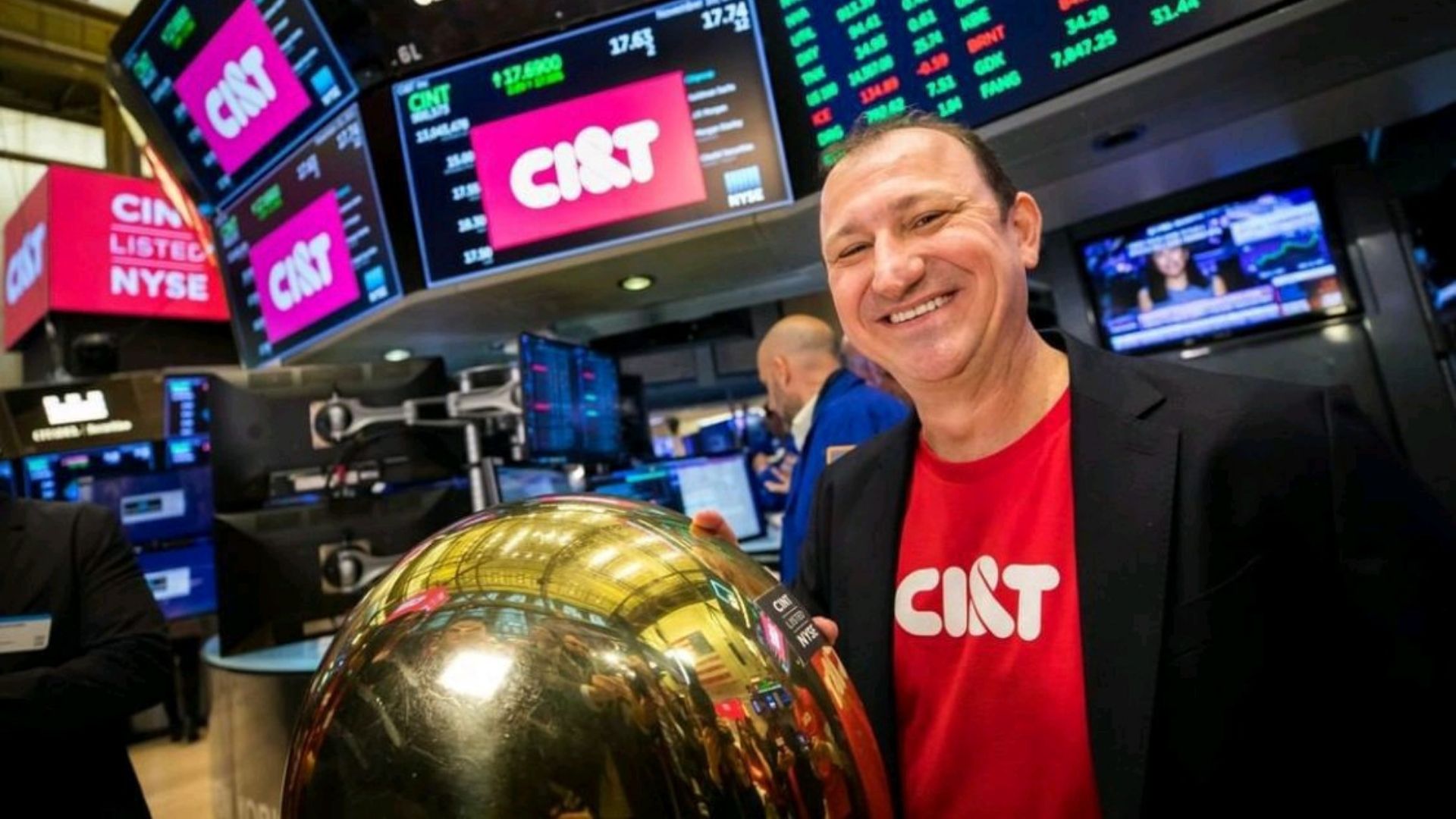 Homem, aproximadamente 40 anos, veste camisa vermelha com terno preto e tem a mão direita sobre um globo dourado. Ao fundo monitores de computador