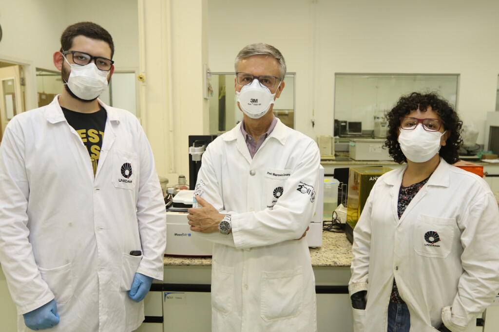 Dois homem e uma mulher posam para a foto. Os três usam óculos, jaleco branco e máscara. Ao fundo o laboratório com banca e equipamentos usados na pesquisa.