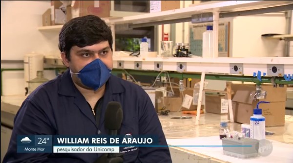 Pesquisador jovem de cabelos escuros, usa máscara e jalecos azuis e segura microfone em entrevista no laboratório