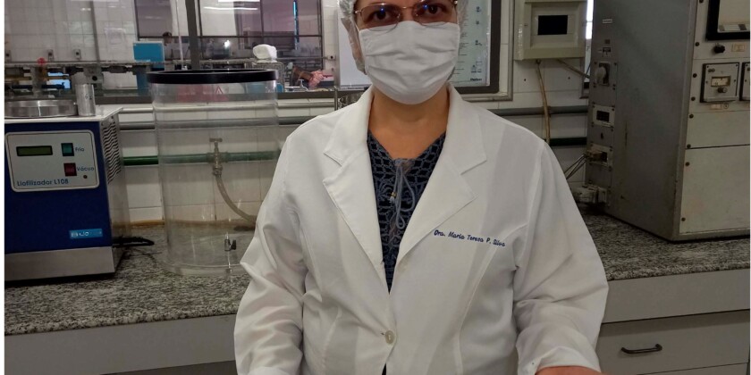Em laboratório, mulher branca de jaleco, touca e máscara segura assadeiras com uma espécie de farinha vegetal