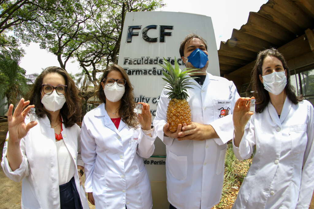 três pesquisadoras seguram frascos com pó branco da bromelina e um pesquisador segura um abacaxi fruta, estão em área externa arborizada com placa da Faculdade de Ciências Farmacêuticas ao fundo