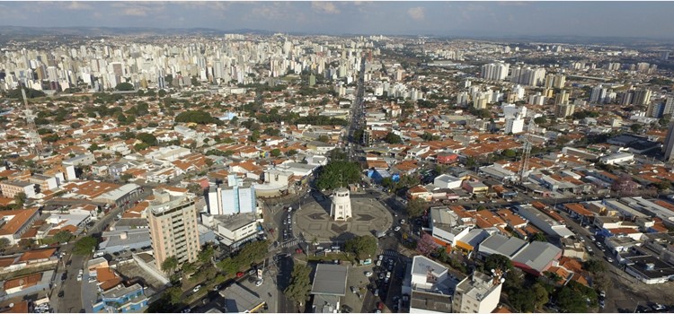 Foto aérea de drone sobrevoando o centro da cidade de Campinas. A foto está sobre uma praça cercada de prédios e casas. O dia está ensolarado com nuvens. Fim de descrição.