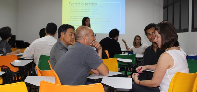 A foto apresenta diversas pessoas divididas em grupos em uma sala com cadeiras universitárias coloridas. No telão ao fundo está escrito "Exercício prático". Fim da descrição.