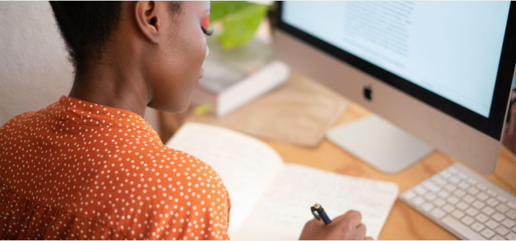 Uma mulher negra está sentada em uma mesa de escritório escrevendo em um caderno pequeno. É possível ver um computador ligado em sua mesa.