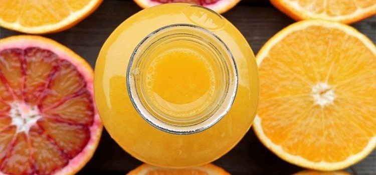 A foto mostra uma jarra de vidro com suco de laranja, vista por cima, ao lado de fatias de laranja cortadas. O fundo da foto é escuro.