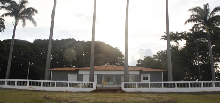 A foto mostra uma casa cinza cercada de um muro baixo branco e de palmeiras. Na frente, um gramado verde e no fundo árvores altas, o tempo está nublado. Fim da descrição.
