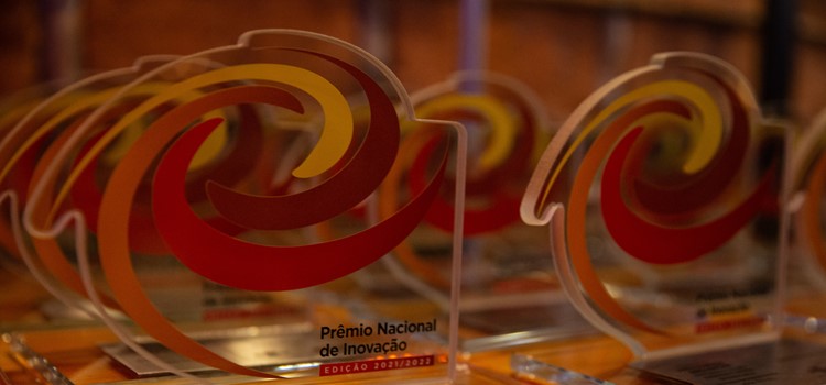 Foto mostra vários troféus em acrílico transparente com desenhos circulares em vermelho e amarelo e escrito Prêmio Nacional de Inovação 2021-2022. Fim da transcrição.