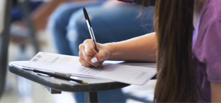 A imagem mostra a mão de uma mulher estudante segurando uma caneta durante a realização de uma prova. Fim da descrição.