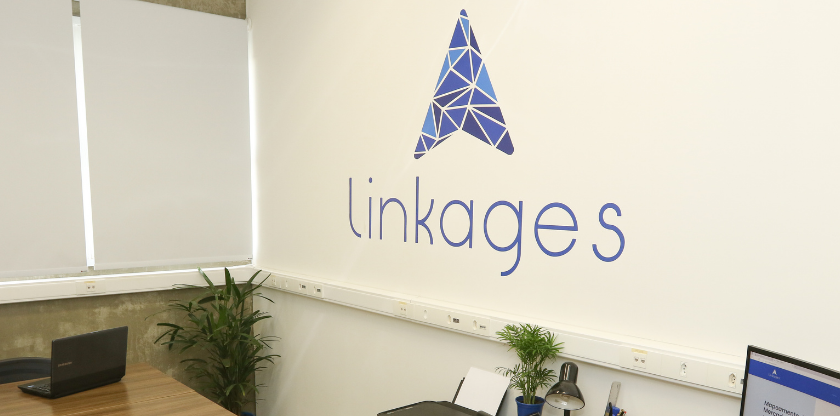 Imagem da sala de escritório da startup no Parque Científico da Unicamp. A imagem foca em uma parede branca com o logo da empresa, chamada Linkages. Fim da descrição.