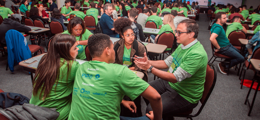 A imagem mostra um grupo de alunos sentado em uma mesa discutindo com um mentor durante uma das edições anteriores do Desafio Unicamp. Fim de descrição.