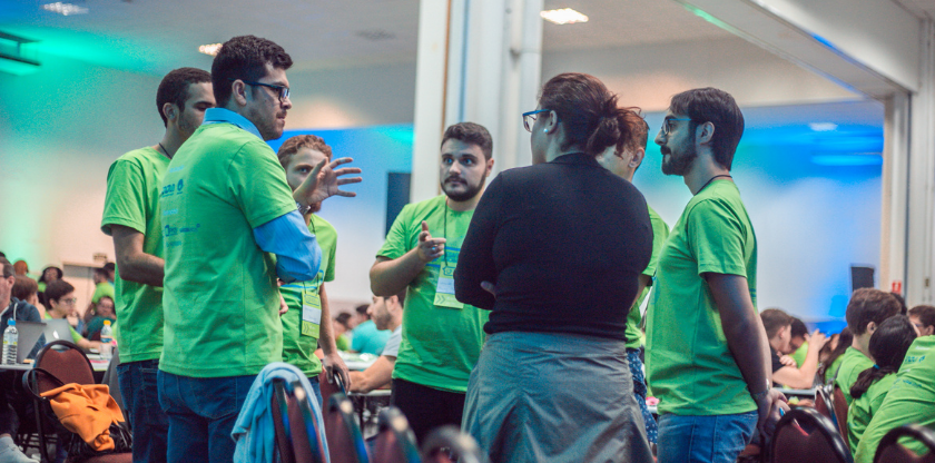 Sete pessoas em pé posicionados em círculo conversando em entres. São 6 meninos vestindo a camiseta do Desafio Unicamp 2019 e uma menina de blusa preta. Todos estavam no Workshop presencial do Desafio Unicamp 2019, organizado pela Inova Unicamp. Fim da descrição.