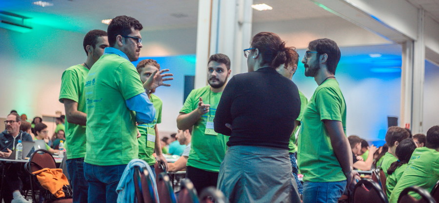 Sete pessoas em pé posicionados em círculo conversando em entres. São 6 meninos vestindo a camiseta do Desafio Unicamp 2019 e uma menina de blusa preta. Todos estavam no Workshop presencial do Desafio Unicamp 2019, organizado pela Inova Unicamp. Fim da descrição.