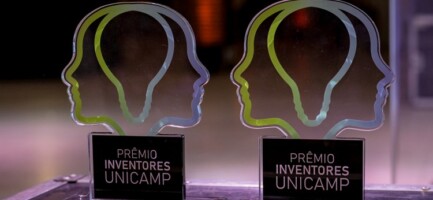 Foto de dois troféus em foco do Prêmio Inventores da Unicamp. O troféu é o logo do Prêmio, que forma uma lâmpada com duas metades de cabeça ao redor. Ele é em degradê do verde ao azul como as cores da Inova Unicamp. Fim da descrição.