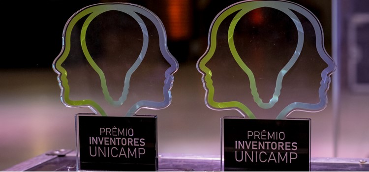 Foto de dois troféus em foco do Prêmio Inventores da Unicamp. O troféu é o logo do Prêmio, que forma uma lâmpada com duas metades de cabeça ao redor. Ele é em degradê do verde ao azul como as cores da Inova Unicamp. Fim da descrição.