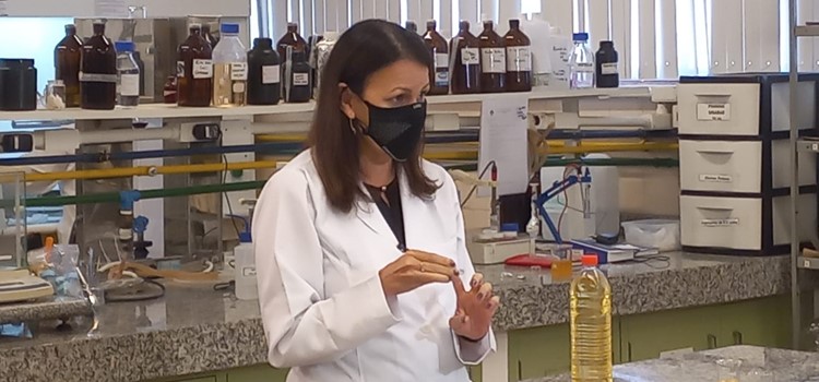 Foto da professora Rosiane, uma mulher branca que veste jaleco branco e máscara preta de proteção, está em laboratório da Unicamp com bancadas de mármore com frascos de vidro. Na frente da pesquisadora há uma garrafa de óleo de linhaça. Fim da transcrição.