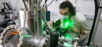 Foto colorida mostra pesquisador homem, branco. Ele mexe em equipamento metálico e tubular que emite numa das extremidades uma luz verde.