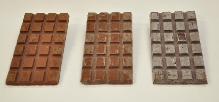 Foto de três barras de chocolate em estágio de embranquecimento diferentes. As barras estão sobre uma mesa branca. Fim da transcrição.