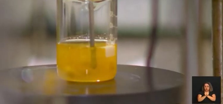 recipiente de vidro transparente com óleo amarelo no interior sendo agitado por equipamento metálico. Fim da descrição.