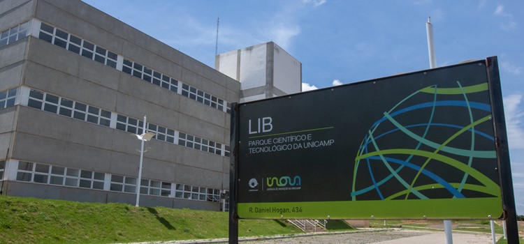 Foto colorida de placa onde está escrito o nome do prédio LIB com logos da Inova e do Parque Coentífico. Ao fundo o prédio cinza com três pavimentos. Fim da descrição.