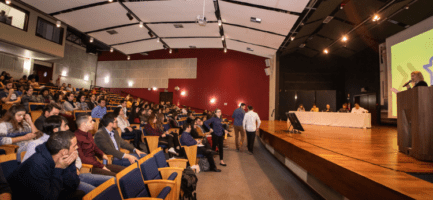 A foto mostra um auditório com pessoas sentadas e um palco com apresentação de participantes da final do Desafio Unicamp 2019. Fim da descrição.