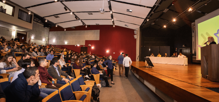 A foto mostra um auditório com pessoas sentadas e um palco com apresentação de participantes da final do Desafio Unicamp 2019. Fim da descrição.