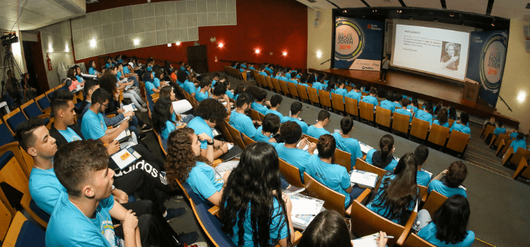 Foto de um auditório com pessoas sentadas em frente a um painel. Imagem tirada no workshop do Inova Jovem 2019. Fim da descrição.
