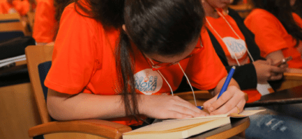 Menina com camiseta laranja, cabelos escuros e presos sentada em uma cadeira escrevendo em um caderno.