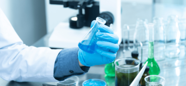 Mão de uma pessoa com luva azul segurando um recipiente com um líquido azul em um laboratório. A imagem é ilustrativa aos exames de autismo. Fim da descrição.