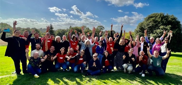 Foto colorida de time da CI&T UK. O grupo com mais de 20 pessoas está em um gramado verde sob um céu azul parcialmente com nuvens em dia ensolarado. Fim da descrição.