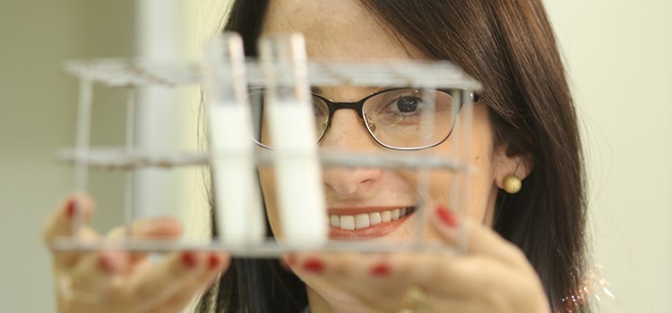 A pesquisadora Ana Badan, uma mulher jovem, branca, de cabelos castanhos, lisos e que usa óculos, segura uma bandeja com amostras de um líquido branco em tubetes de vídeo. A foto foca nas mãos e no rosto da pesquisadora. Fim da descrição.