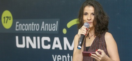 Foto de uma mulher branca, de cabelo castanho curto ondulado com microfone na mão. Fundo azul escuro escrito 17º Encontro Anual Unicamp Ventures. Fim da descrição