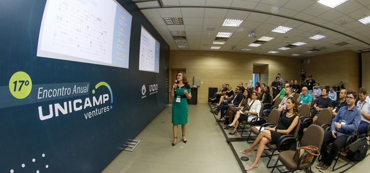 Foto lateral de auditório onde público assiste a apresentação da professora Ana Frattini, uma mulher branca de vestido verde. Fim da descrição.